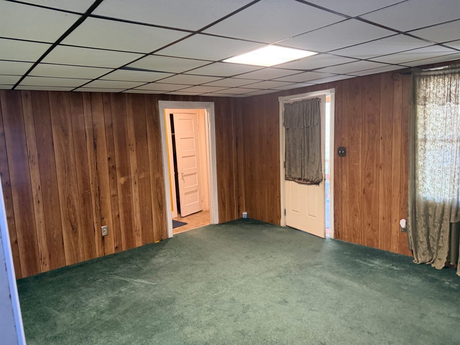 Living room before renovation with wood paneling, drop ceiling and worn green carpet